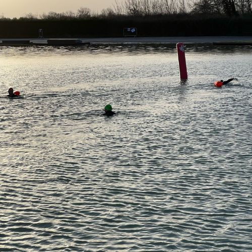 Swimmers in wetsuits and bright swim caps preparing for a cold plunge on New Year’s morning in the Cotswolds.