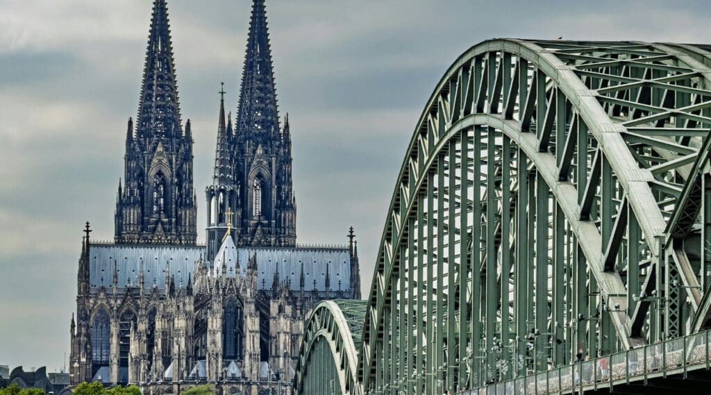 The Hohenzollern Bridge steel arch spanning the Rhine with Cologne Cathedral's twin Gothic spires rising dramatically behind it