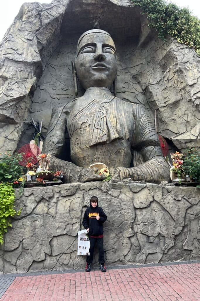 Dylan standing in front of the giant Asia-themed deity statue carved into rock at Phantasialand, with red parasols and floral offerings at its base