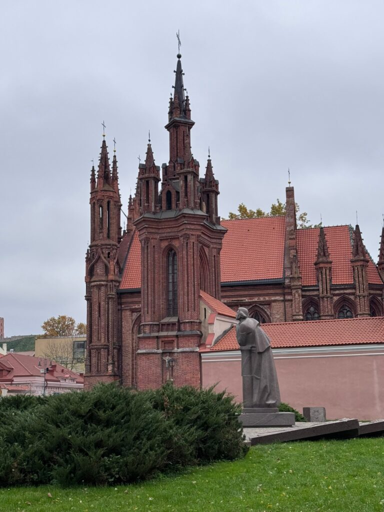 Red-brick Gothic church with pointed spires and a stone statue in the foreground in Vilnius